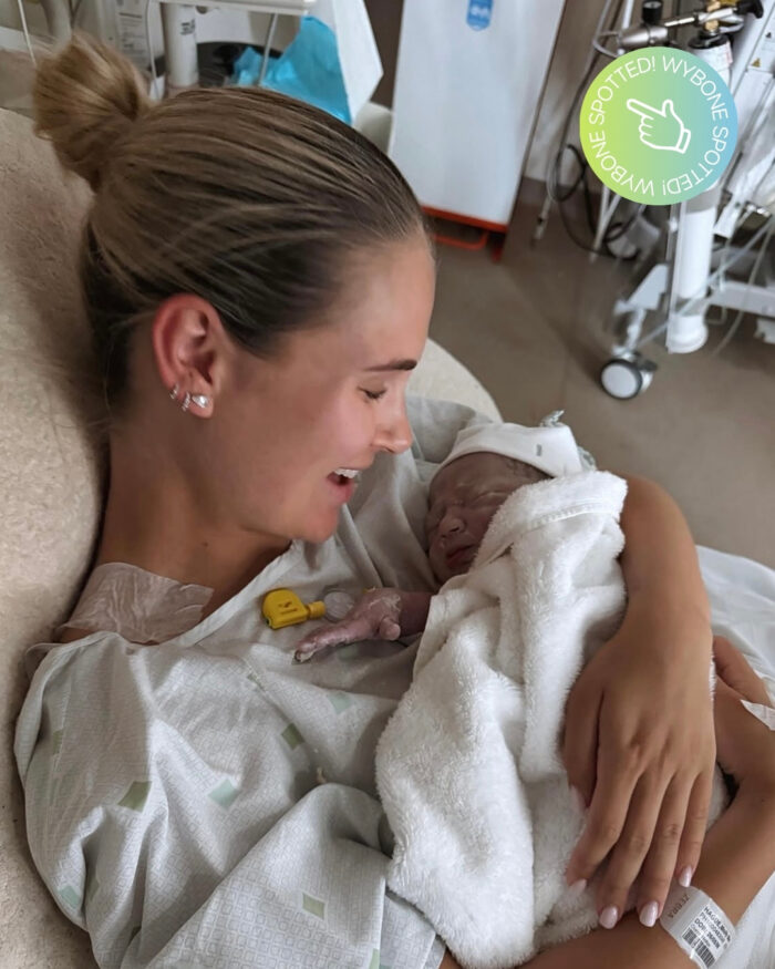 Molly-Mae Hague holding her newborn baby in a hospital room shortly after birth. Medical equipment is visible in the background, including a Wybone clinical waste sack holder beside the hospital bed.