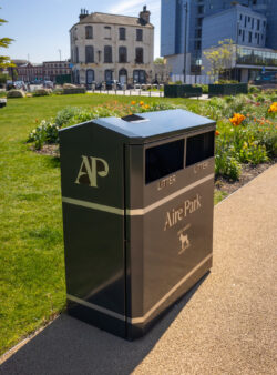 Wybone MLB/224 dual litter and dog waste bin with bespoke Aire Park branding installed beside a landscaped pathway in Aire Park, Leeds. The dark green galvanised steel bin stands among colourful flower beds and green lawns, with modern office buildings in the background on a sunny day.