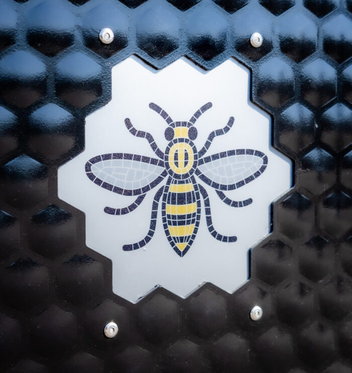 Detailed view of the Manchester Bee emblem on a bespoke Wybone MLB/112 litter bin with a black honeycomb finish. Custom-designed street furniture manufactured for Manchester City Council as part of the city's iconic public realm branding.