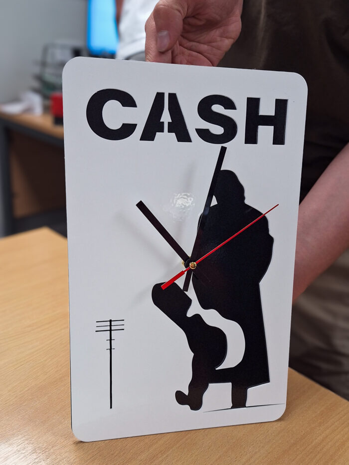Finished laser-cut steel Johnny Cash-themed clock designed by a Year 10 work experience student at Wybone. The bespoke clock features a silhouette of Johnny Cash holding a guitar, the word "CASH" across the top, and black clock hands with a red second hand, showcasing the student's CAD design brought to life through UK steel manufacturing.